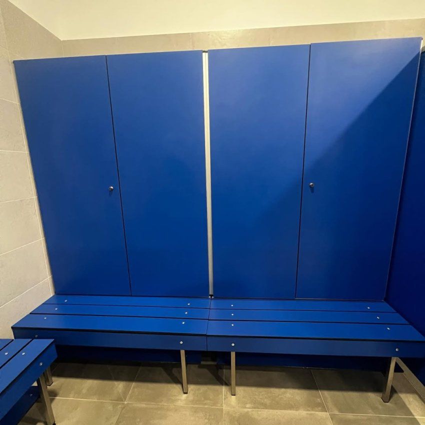 Vestuario con bancos fenólicos azules y cabinas sanitarias del mismo color instaladas sobre suelo cerámico gris