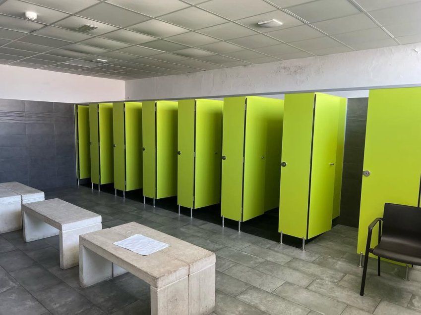 Vestuario deportivo con cabinas fenólicas verdes, bancos de hormigón y suelo cerámico gris en instalación de uso colectivo