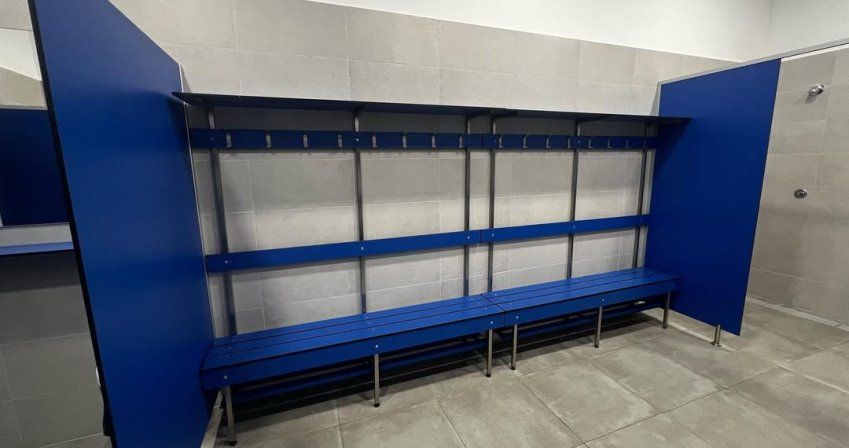 Banco fenólico azul con percheros integrados en vestuario deportivo con paredes y suelo cerámico gris