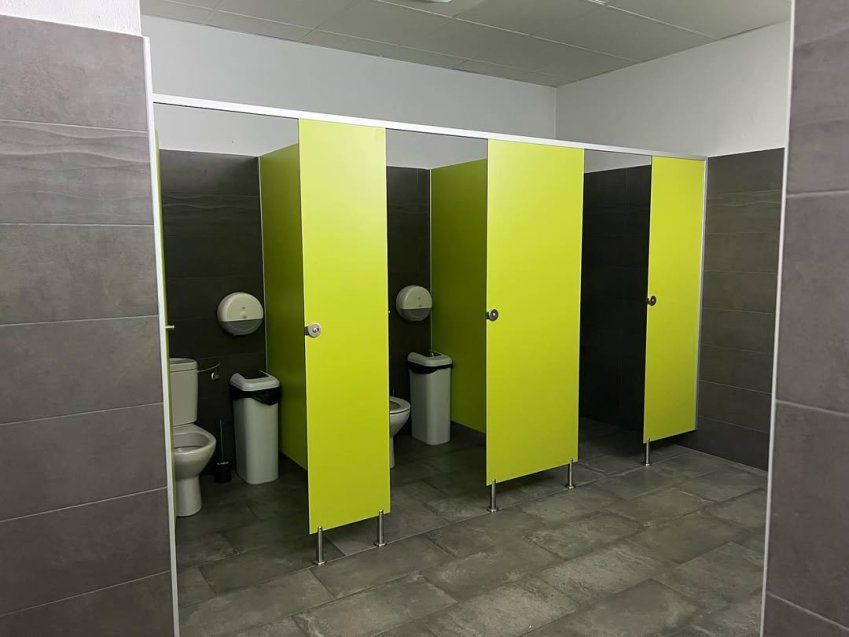 Cabinas fenólicas verdes instaladas en aseo público con puertas compactas, suelo gris y separadores para vestuarios y baños colectivos